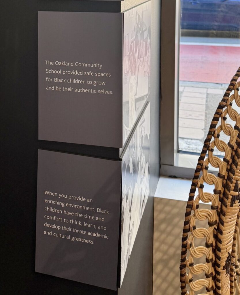 Words on a silver pillar tell about Oakland Community School next to a wicker chair and before a large window.