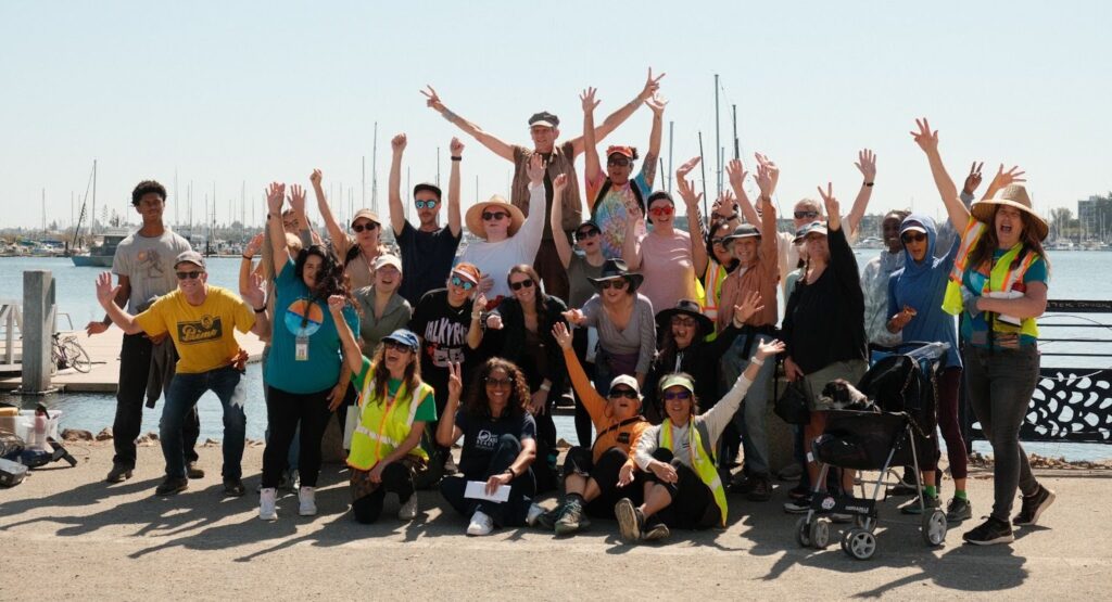 About 35 people, some seated on the ground, others standing, about 3 rows deep raise hands and fists in celebration in front of a body of water.