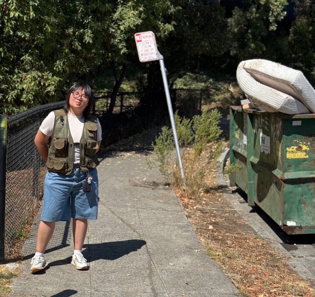 A woman in short dark hair and glasses, wearing a flack vest over a white T-shirt and long jeans shorts stands on a sidewalk beside a fence to her right. To her left is a green dumpster with a mattress poking out of the top.