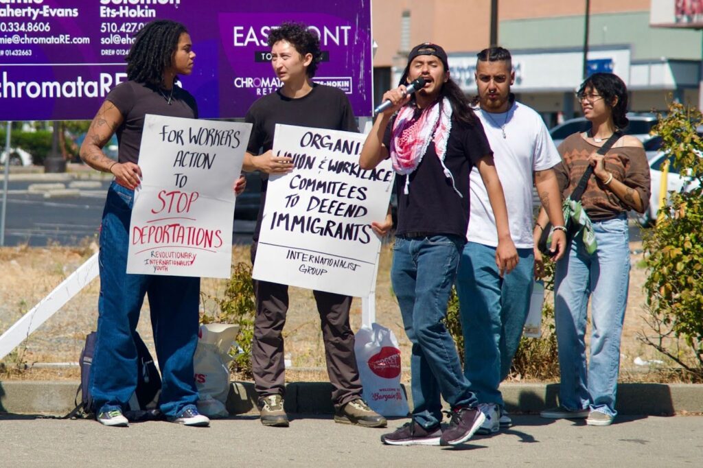 Five people stand in a row on a stretch of sidewalk in front of a building. The two on the left are holding signs, the one in the middle is holding a microphone, and the two on the right are looking at the others.