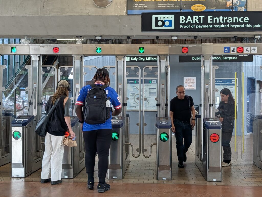 Five fare gates in a subway station are at the center, with two women lined up to go through the second gate, which has a green arrow, a man coming forward through a gate with a red circle and a woman in the next gate tapping her card.