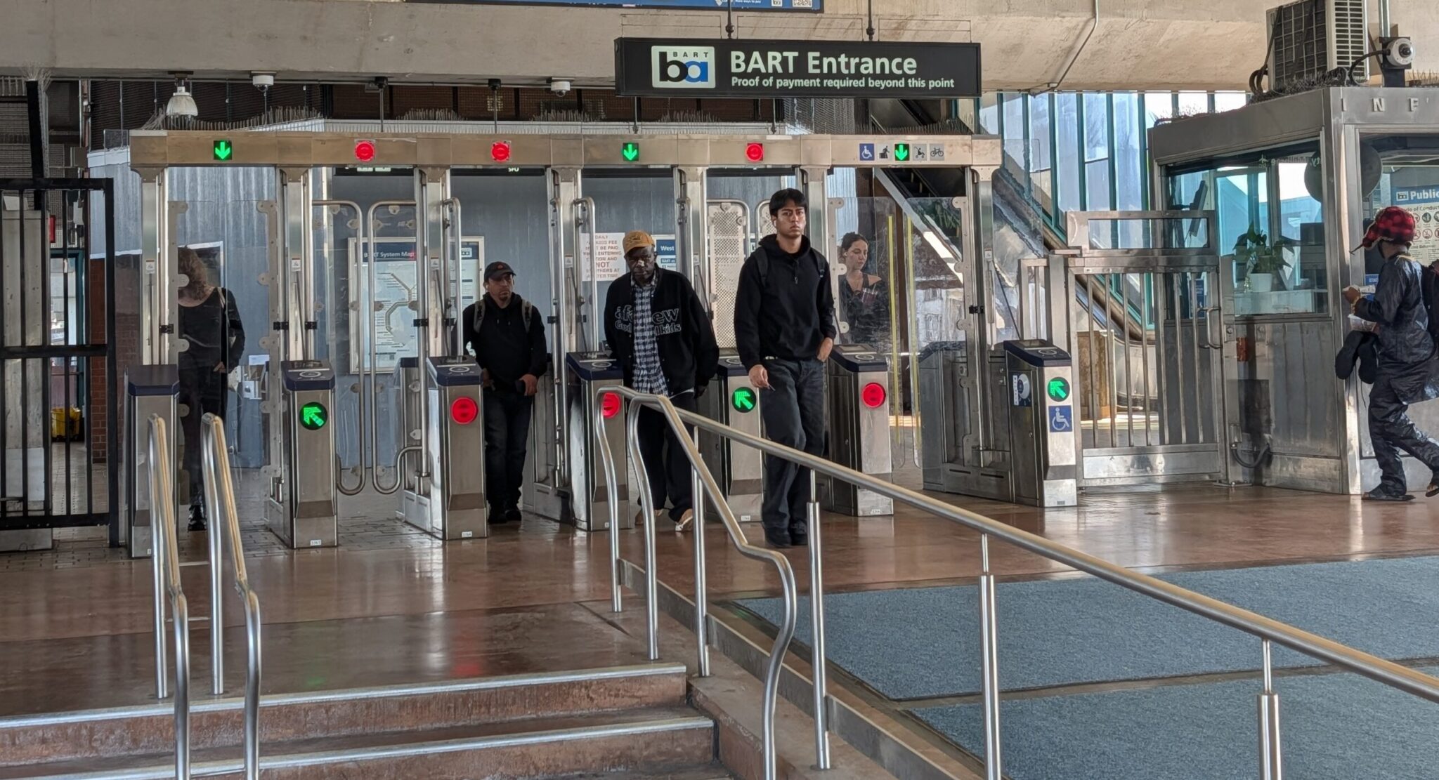 Six subway entrance gates are at the center of this picture inside a subway station. The first on the left has a lit green arrow, followed by two red lit circles, then green arrow, red circle, green arrow. Six people are in various stages of going through the gates.