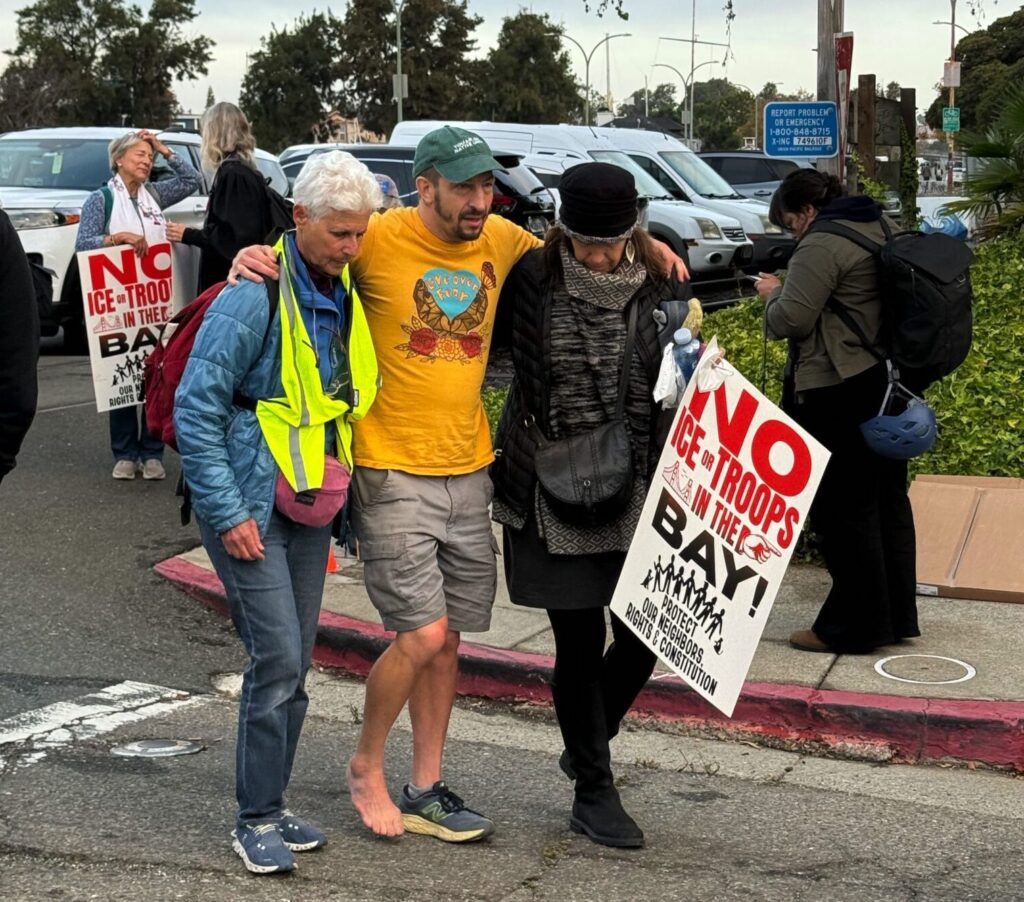 A man hobbles with one sneaker on and a barefoot, supported by two women in a street.