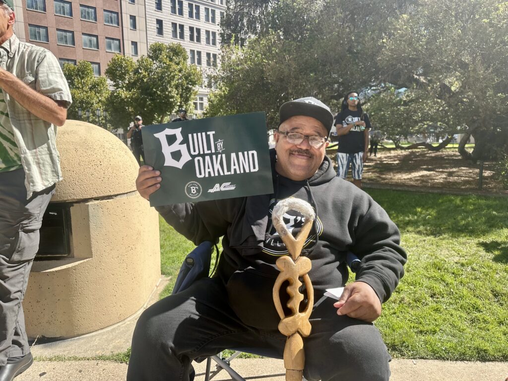 A man sits in a folding chair in a park holding a "Built in Oakland" sign. He is dressed in black pants and black hoodie. In front of him is an ornate, whittle wooden stick that may be a cane.