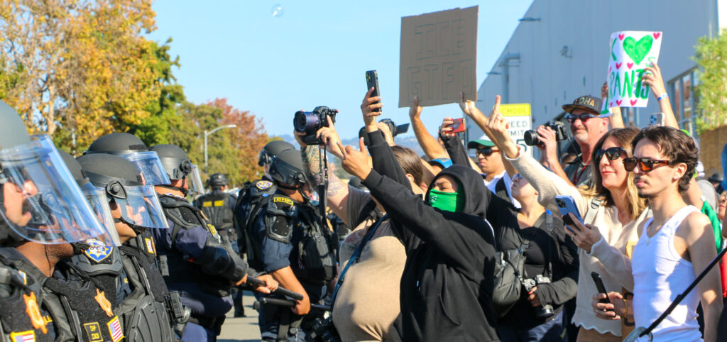 Protesters on the right hold phones and cameras up as they facedown officers in riot gear.