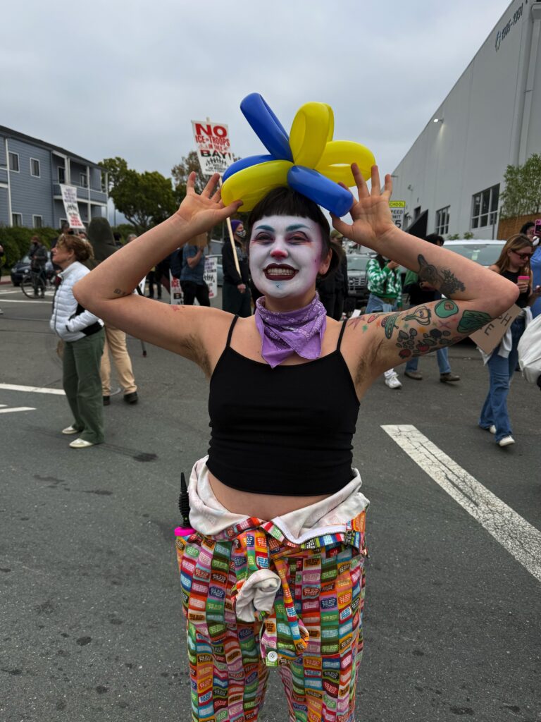 A woman in a white clown face and red nose stands in a street with blue and yellow balloons on her head.