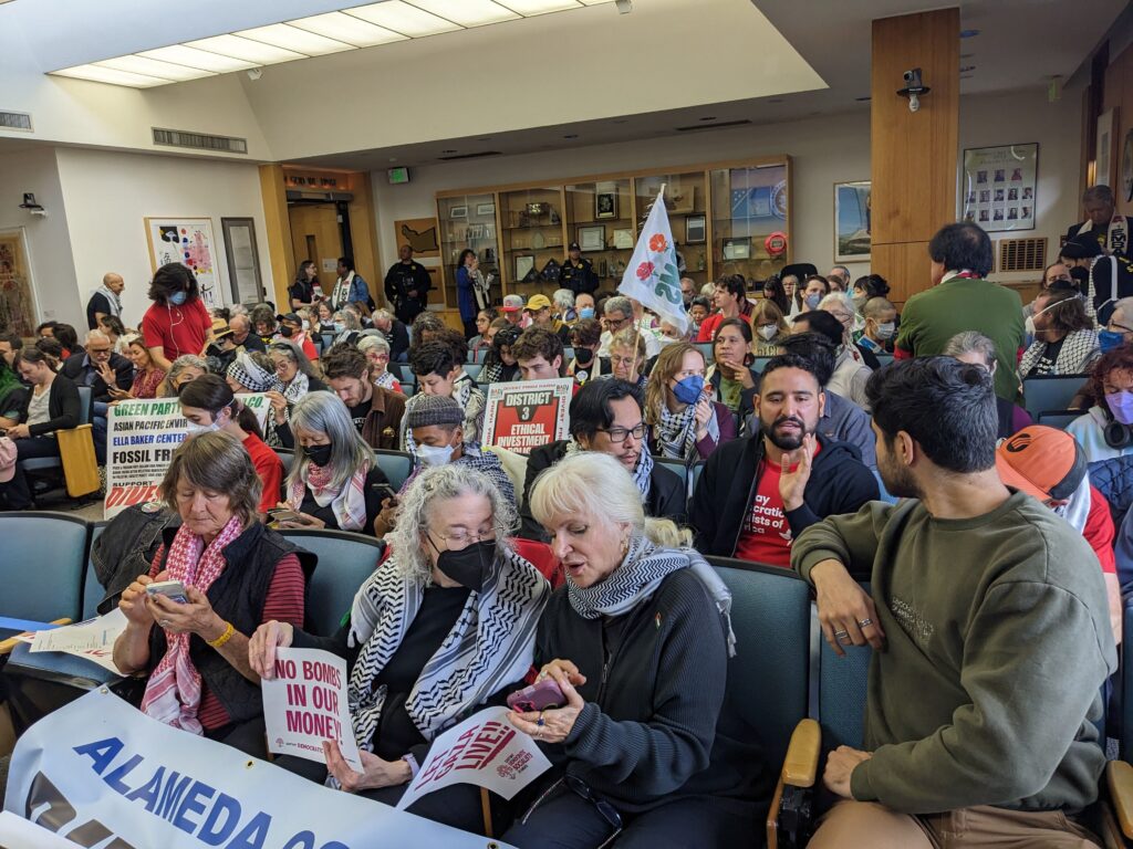 People are packed into a meeting room, the three women in the first row looking at their phones, people in back rows holding signs, a police officer stands by the door in the rear.