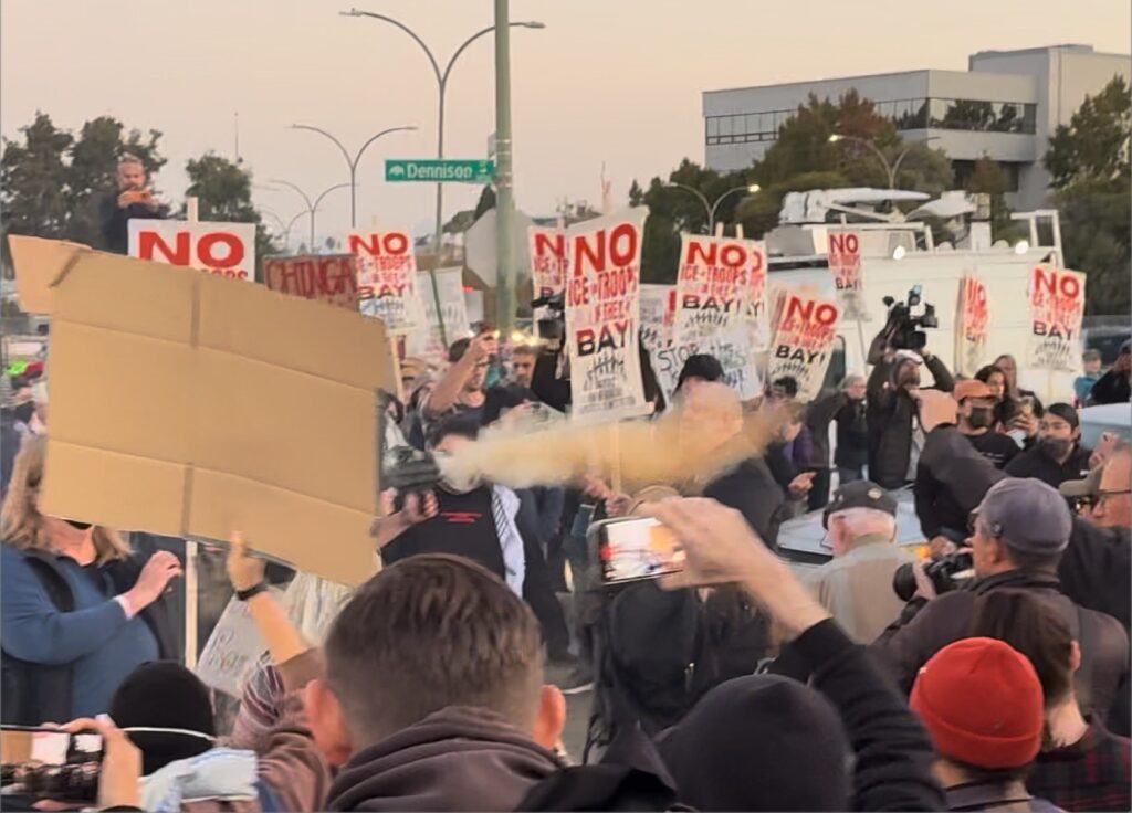 A flash of yellow smoke is released in the middle of a protest with people standing in a circle holding signs on an Oakland street.