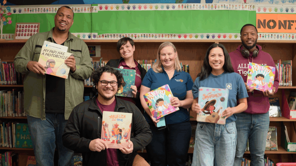 Six adults pose for the camera holding children's books in a classroom standing in front of a long book shelf.