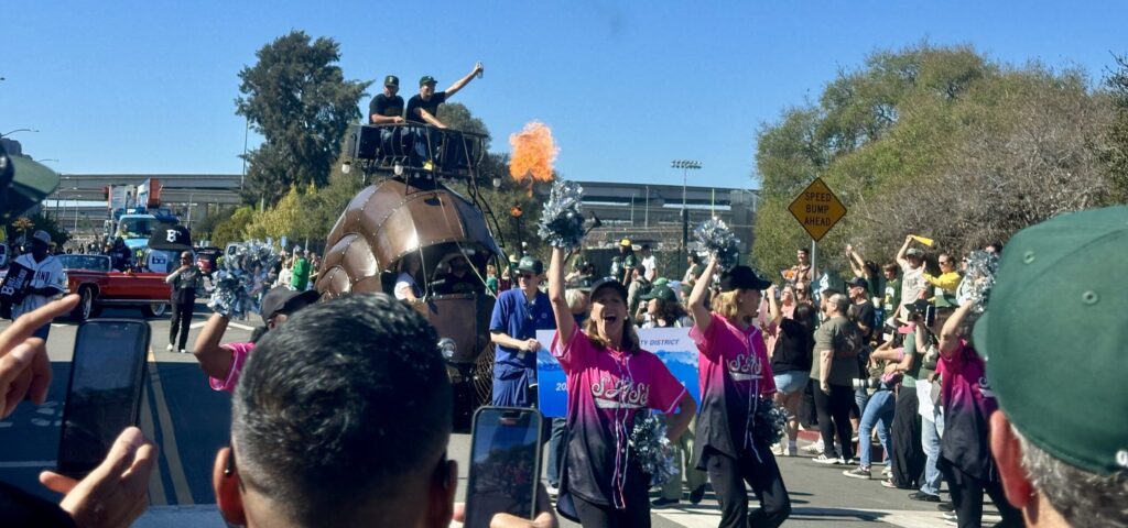 A crowd lines the path in a park as a brown barrel-like vehicle with two guys sitting atop it in a basket wheels by. In the foreground, women in pink T-shirts and pompoms cheer.