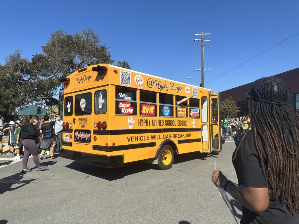 A small yellow school bus is in the center of the screen. It has stickers all over the windows, mostly with words on them. An arm is sticking out of the second of five windows on the right side. In the foreground, a woman with long dark braids stands with her back to the camera. In the rear, a crowd of people is slightly visible.