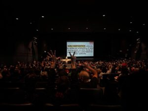 In a dark auditorium, a white screen is lit with words about non cooperation. We see the backs of the full audience, with someone in the foreground holding up five fingers in their left hand and two in their right.