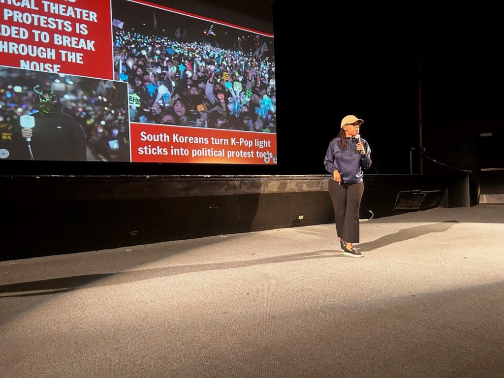 A woman in a blue long-sleeved shirt, black pants and black sneakers, dark braids falls from the back of a tan cap, and glasses, talks into a microphone on a large empty stage. Behind her on a movie screen are images from a South Korean political protest.