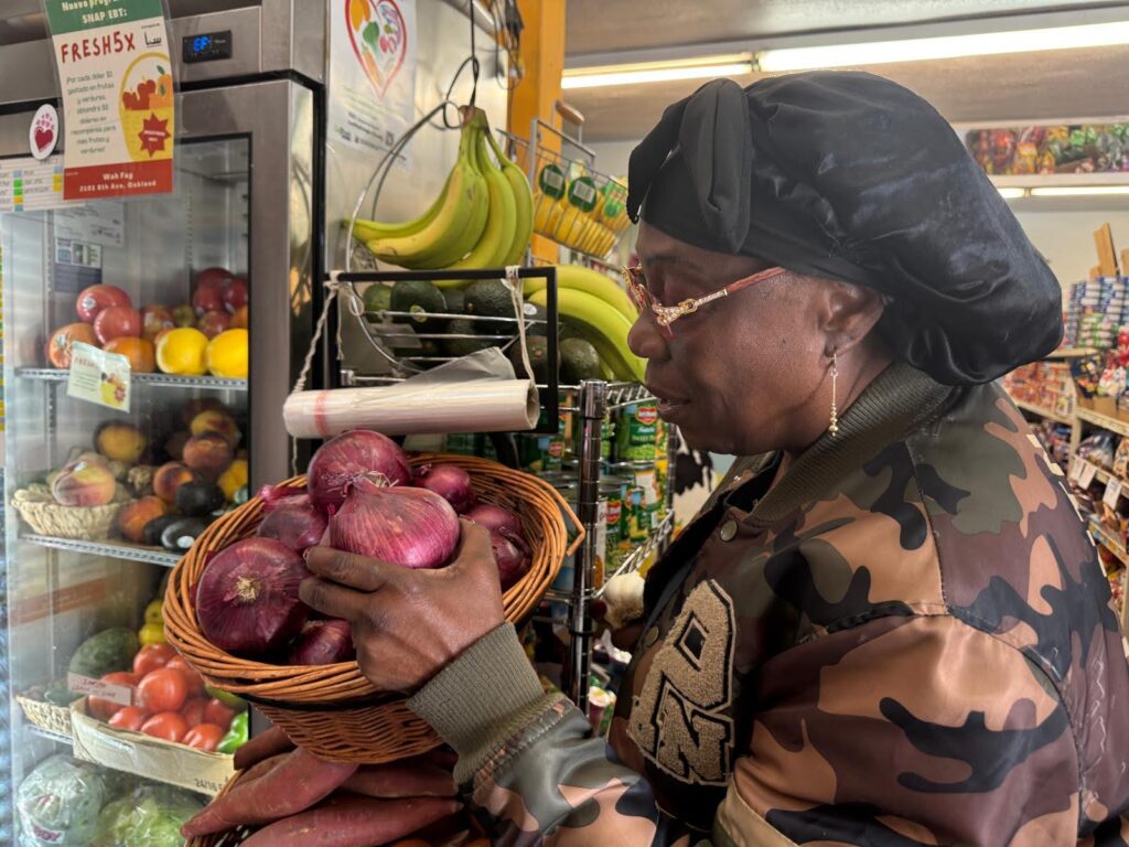 A woman with her hair covered in a black kerchief is in a produce section of a small store, with her left hand on a red onion.