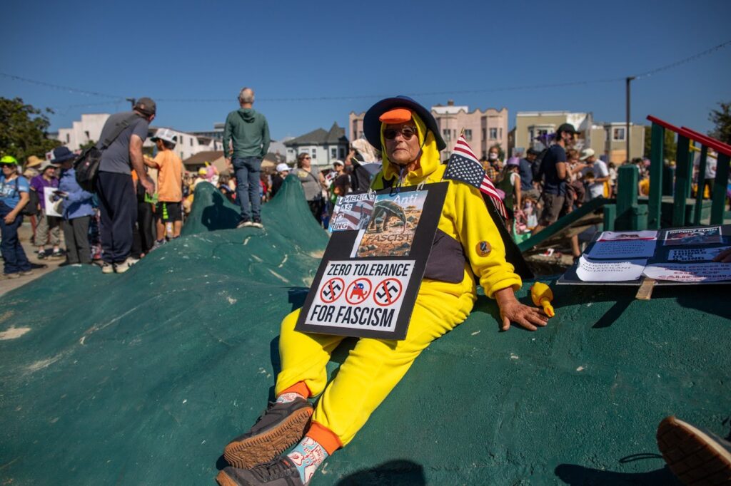 A woman dressed aaas a chick in a yellow jumpsuit with an orange beak, sits atop a green tarp with a sign around her neck and an upside down American flag on her left shoulder.