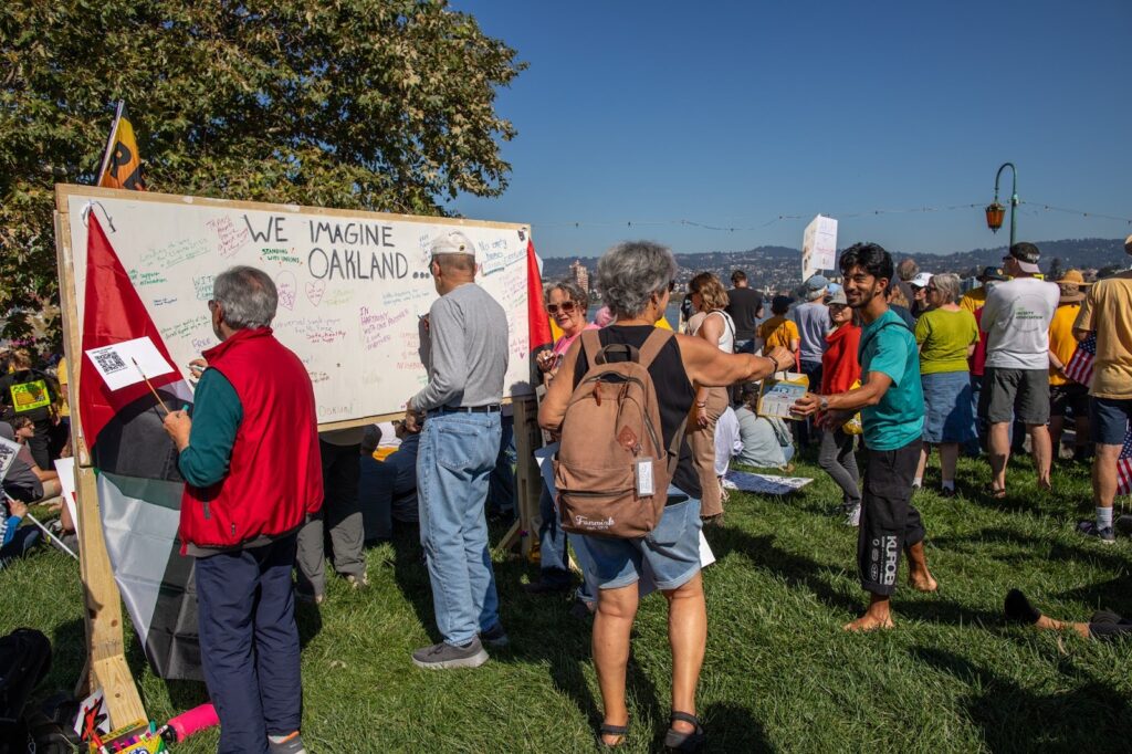 In a park, under a blue sky, dozens of ppeoplel mill about, several standing before a large white board with scribblings under the banner "We Imagine Oakland."
