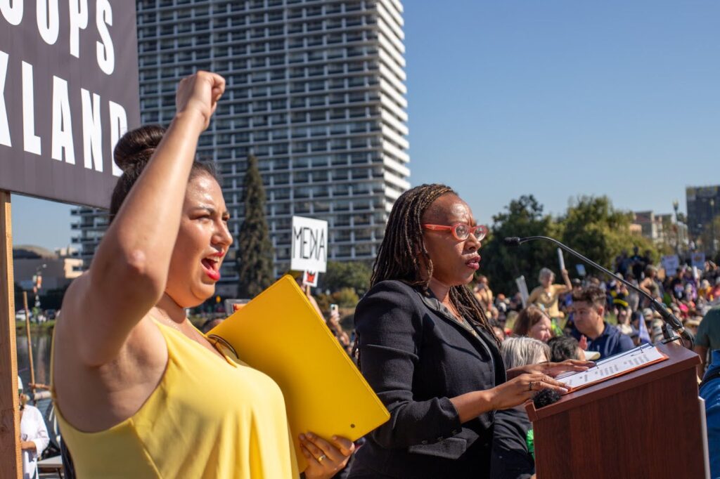 A woman in long dark braids, dressed in a black suit, wearing red-framed glasses stands at a podium at a rally outside where many people are gathered. In the foreground, a woman in a yellow blouse raises her right fist in the air.