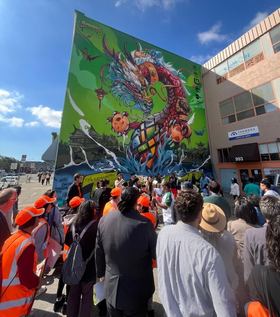 Dozens of people stand beside a wall with a large mural of a dragon in primary colors rising out of the ocean.