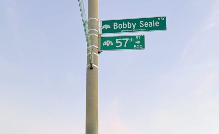Affixed to a pole are two green street signs with tree logos. The one with the words "Bobby Seale Way" sits just above the one that says 57th St.