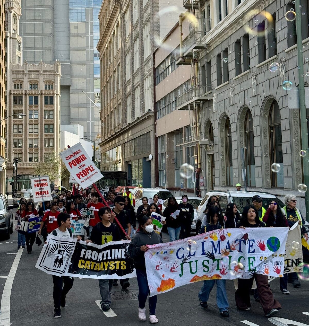 On a narrow street with tall buildings on both sides, a couple dozen teenagers march down the street, carrying signs and banners.
