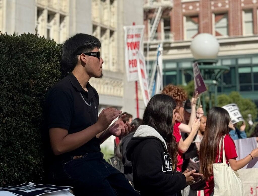 A young man in the focus of the frame is wearing dark clothes and dark sunglasses and is in mid-clap with other teens, who are holding signs and standing outside a white gothic building.