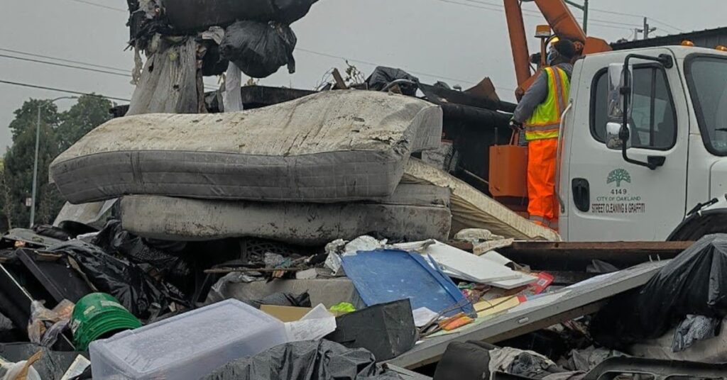 Two dirty mattresses top a pile of assorted trash that includes bins, buckets, black trashbags and wood. A worker in neon orange and yellow clothes stands outside a truck's cab, watching as a claw lifts the trash.