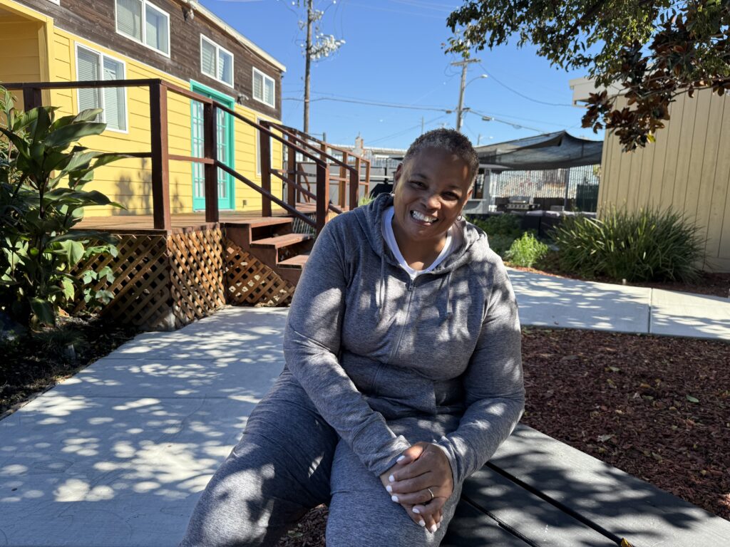 A woman dressed in gray sweatpants and a gray hooded sweatshirt, hair hair cropped and gray, sits outside on a bench smiling.