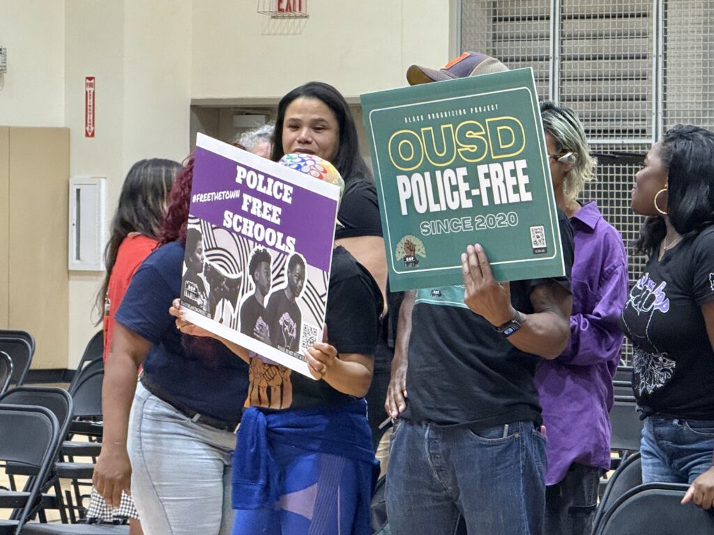 Two people at the forefront holding signs in a room where there are several others behind them.