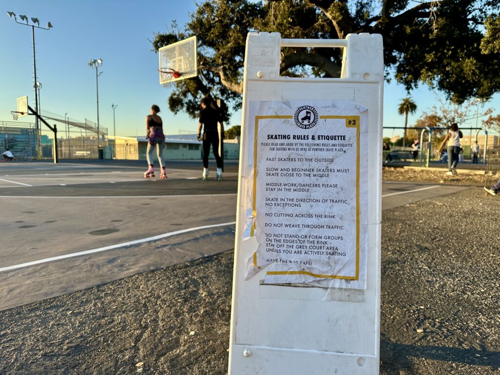 A white sandwich board is in the front of the frame, with a flyer on it about skating rules. It sits on the edge of a basketball court that a couple of people are roller skating on.