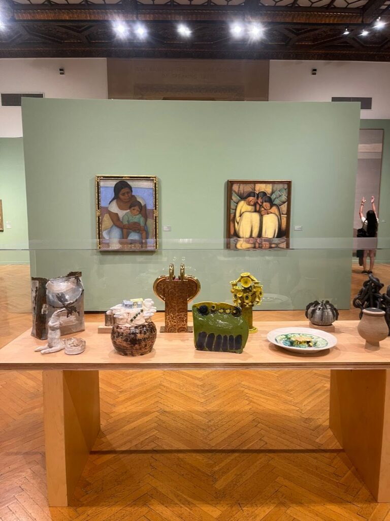 A table is in the foreground with plates and other small art sculptures. Behind it hand paintings by Alfredo Ramos Martinez and Diego Rivera.