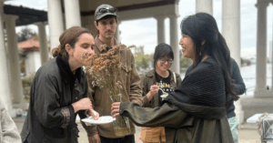 A woman with brown hair tied in a low bun, wearing a black coat, holds a plate while smelling yarrows - a brownish yellowish flower. The flowers are held by another woman wearing a dark green jacket with a shawl around her. Her hair is black and in a semi-ponytail.