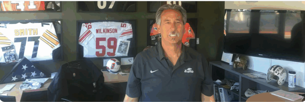 A man in short gray hair and mustache with a short-sleeved navy Nike shirt with a white swoosh over the right breast stands in front of a wall of football jerseys from various teams.