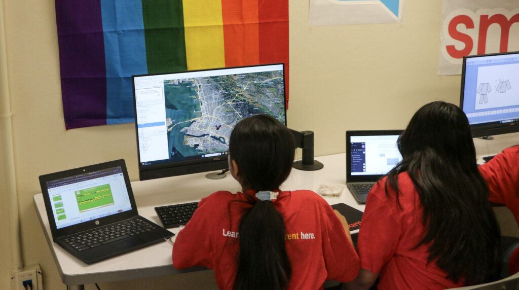 Two young girls, look at a large computer monitor at a table with two laptops on it. They are wearing red T-shirts and both have long dark hair. The one on the left has hers in a pony tail.