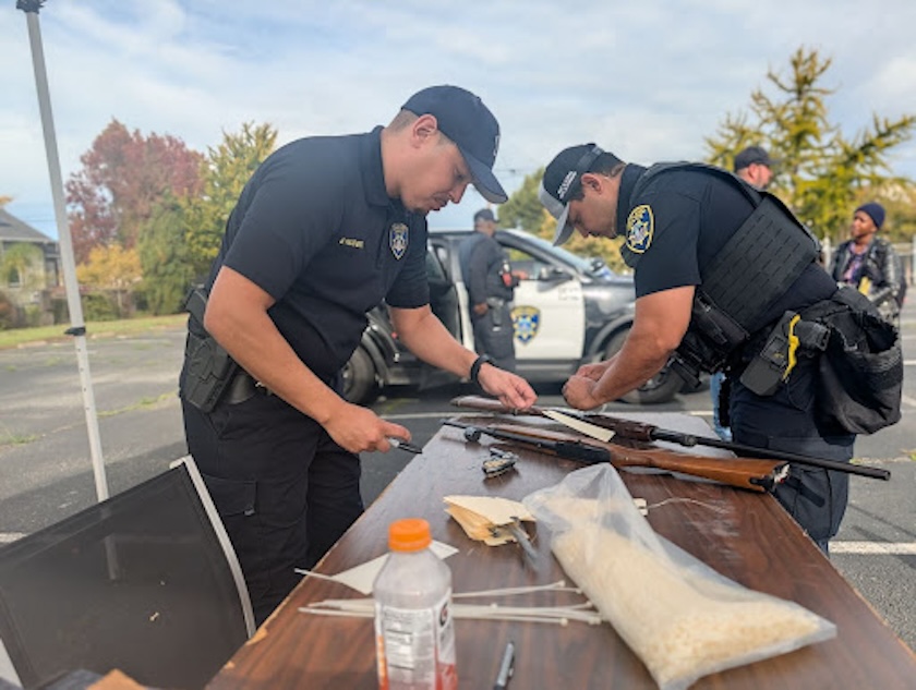 Two police officers label rifles on a wooden table.