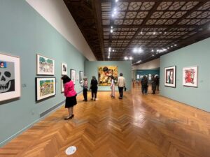 About eight people stand on a hard-wood floor in a museum, looking at large paintings.