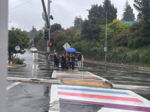 A rainbow stripe runs the width of a wet street, with half a dozen people with umbrellas standing on the far side.