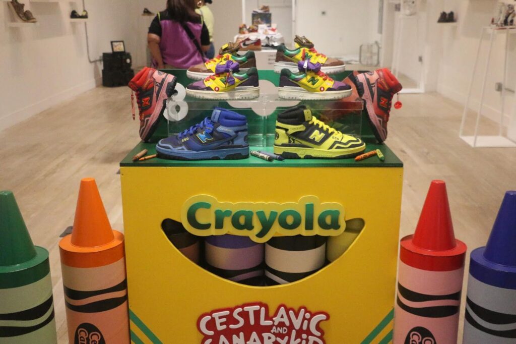A giant box of crayola crayons forms a shelf upon which are high-top sneakers in assorted primary colors.