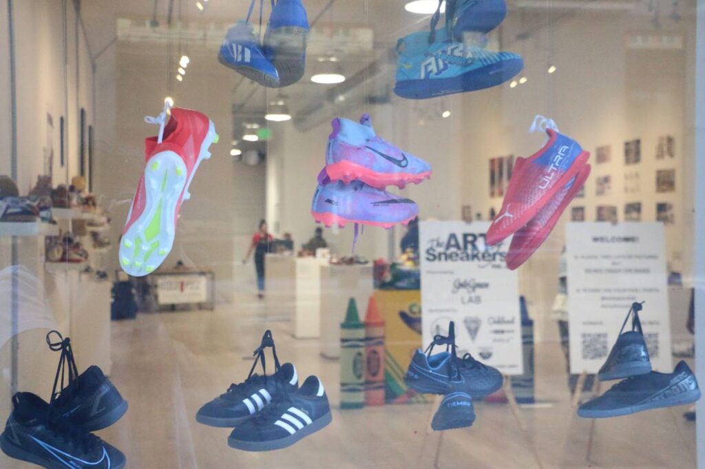 In a storefront window, dangling from suction cups are nine pairs of cleated sneakers in shades of blue and pink.