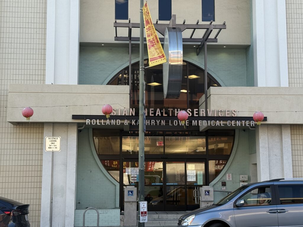 The facade of a building made of beige and white stone with a metal latice and the words Asian Health Services