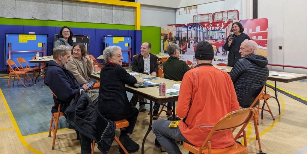 Eight people sit around a long table in a gym, smiling, with a woman in front holding a microphone.