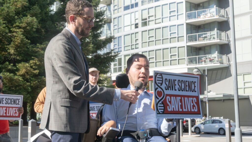 A man in a wheelchair is speaking into a microphone that is being end by a standing man with glasses and a suit jacket, on a sidewalk across the street from a tall apartment building.