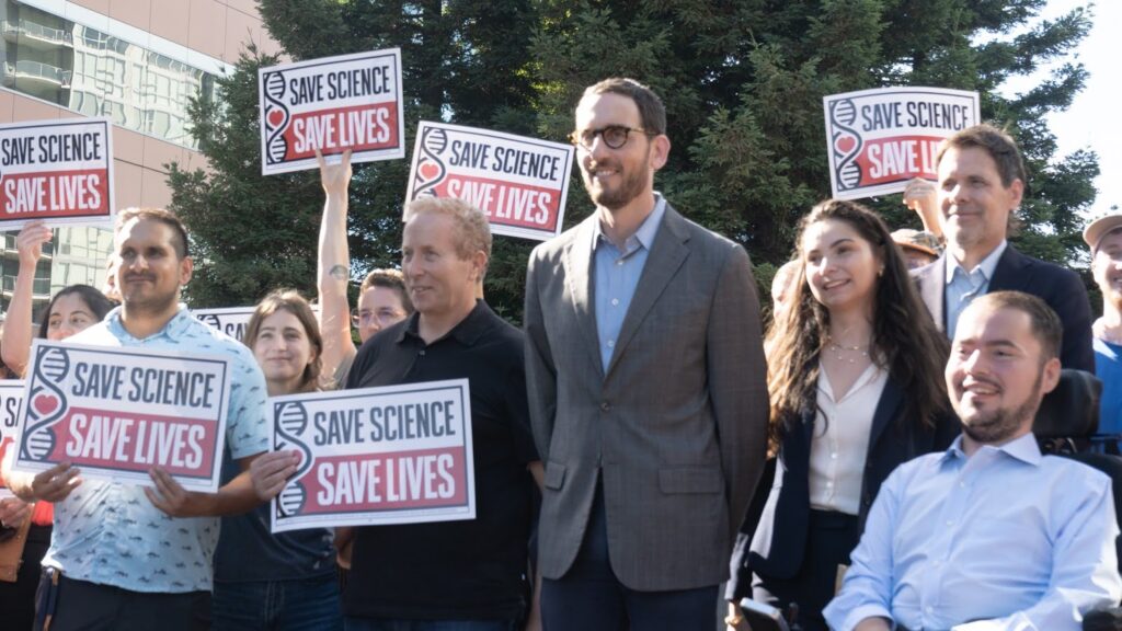 A tall man in glasses and a suit jacket stands in the middle of a group of about a dozen, most of them waving signs.