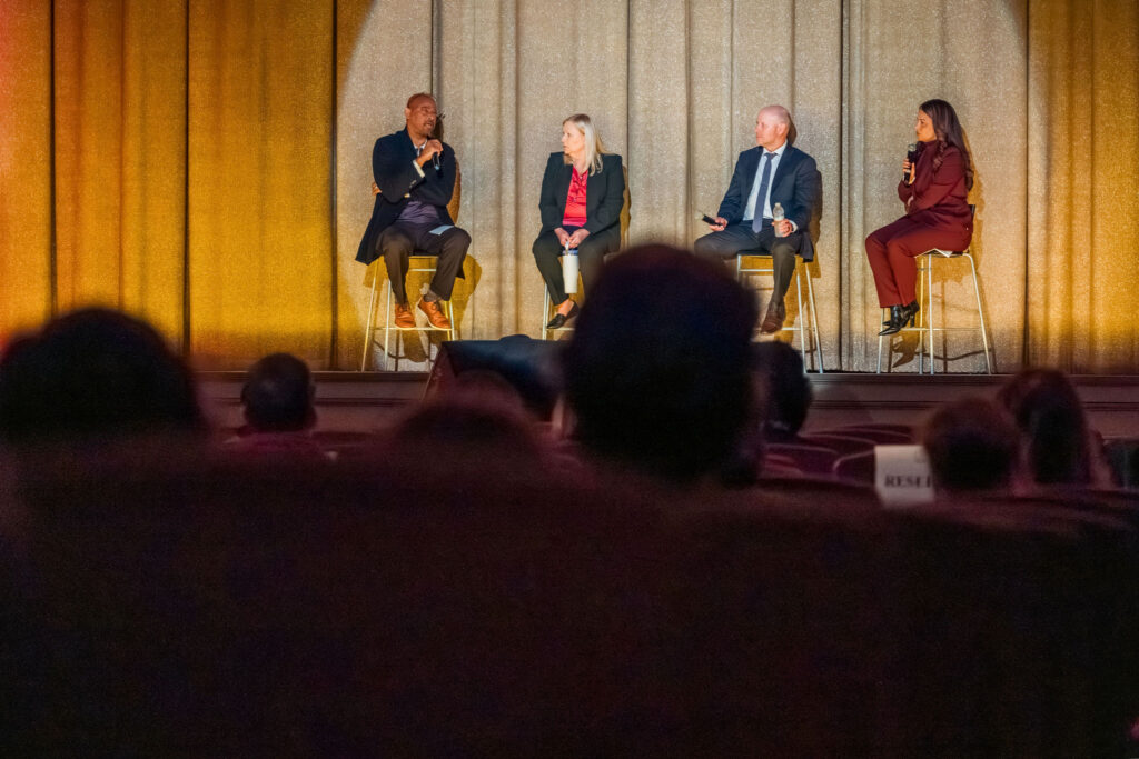 Four people, a man, woman, man, woman, sit on stools on a stage before a dark movie theater filled with people.