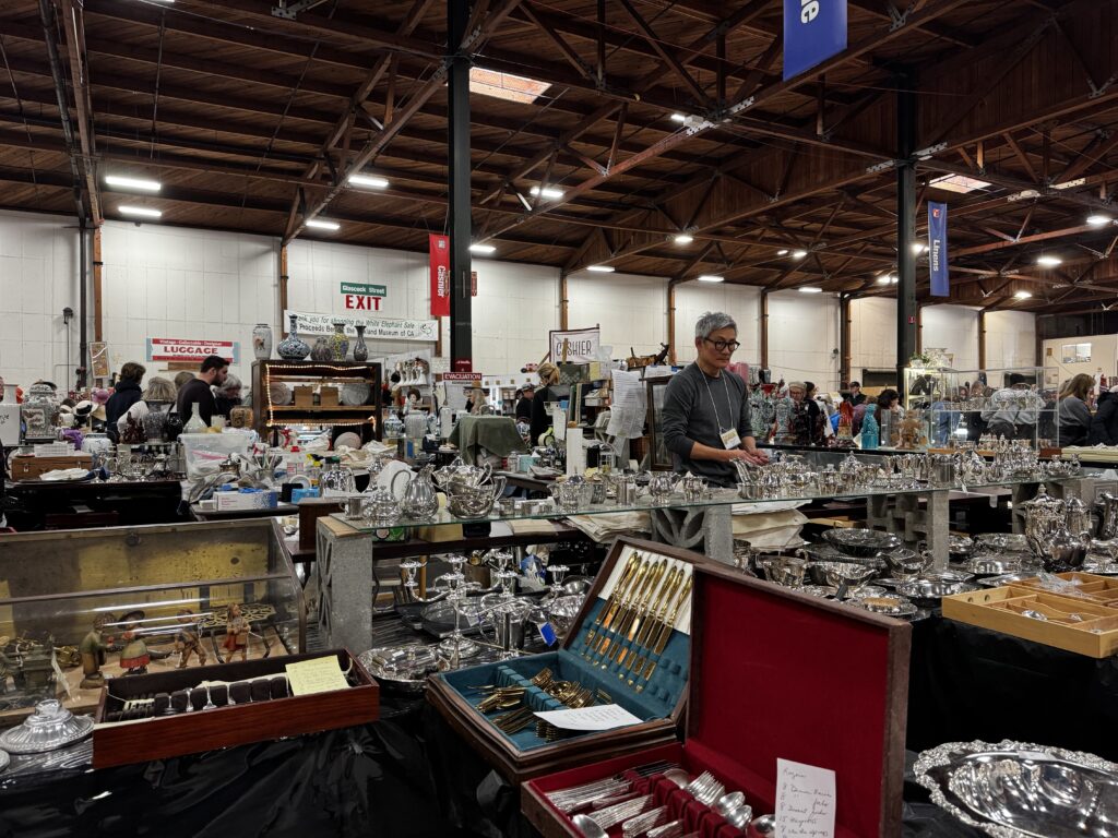In a cavernous room with wooden beamed ceiling, silver and gold platters and cutlery are displayed on glass cases that pack the room. A few people are looking over the objects,