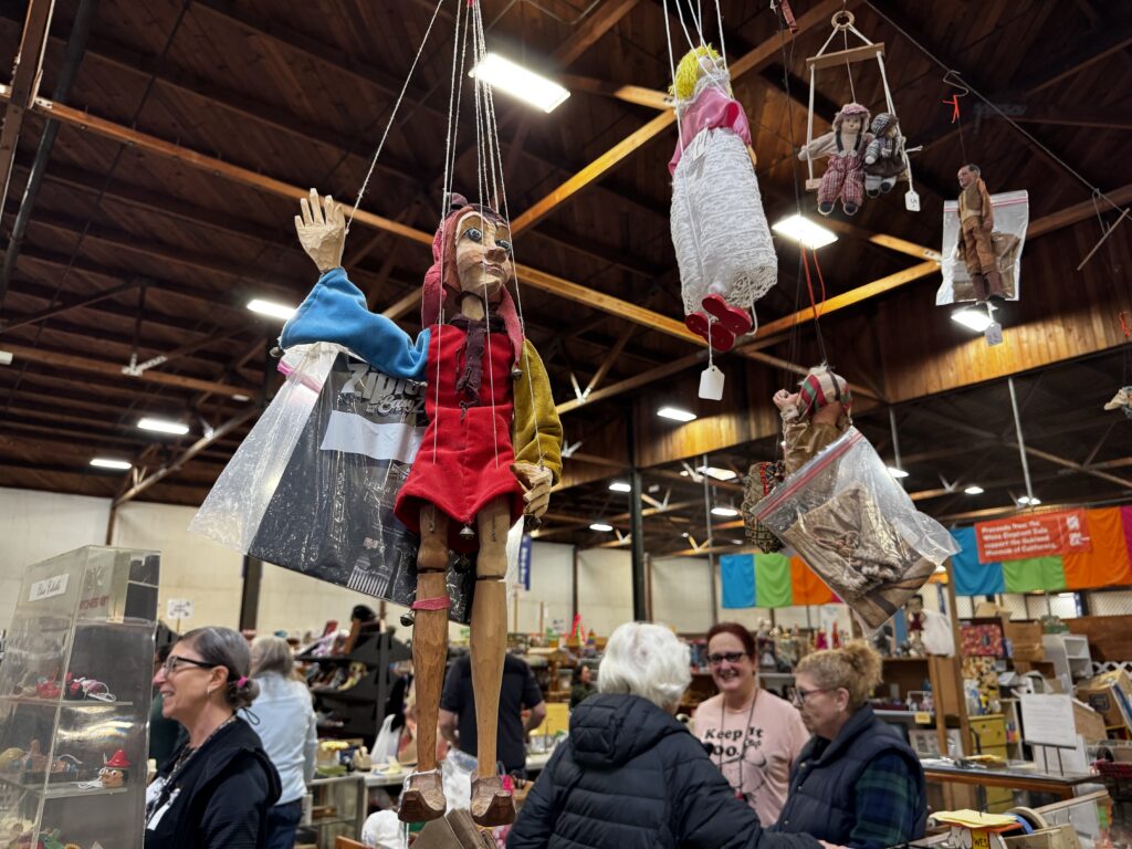 Five marionettes, including a prominent wooden one wearing a red dress hang from rafters in a cavernous room over three women who are having a conversation.