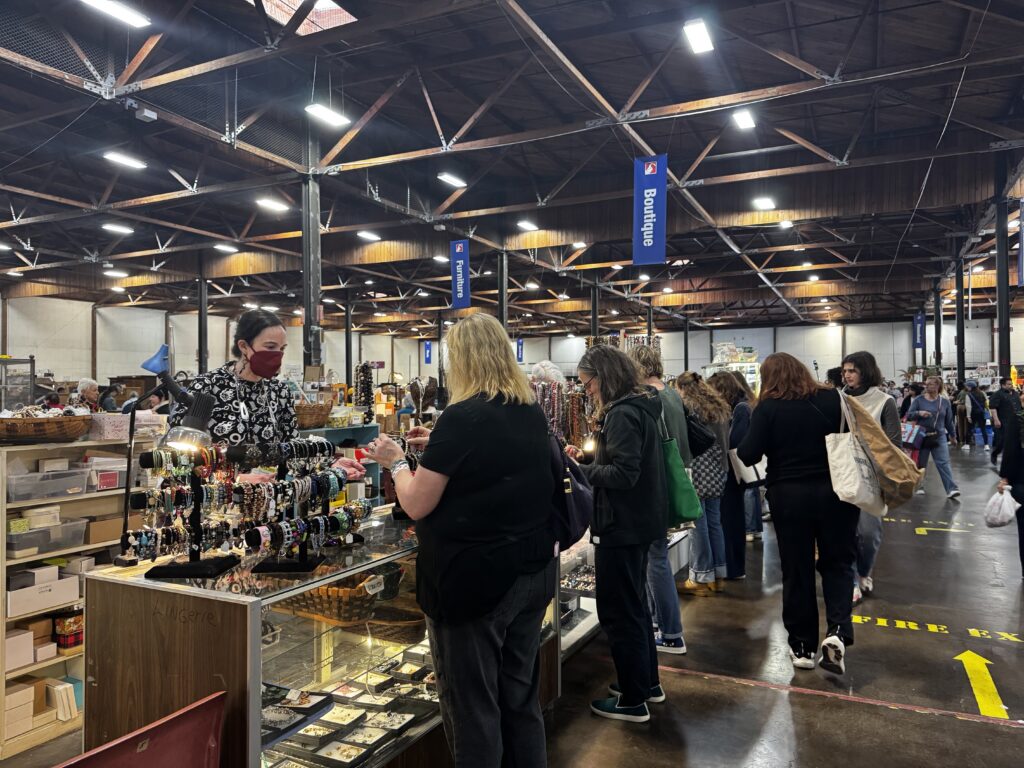 Dozens of woman are crowded around cases of jewelry in a cavernous room with a concrete floor and wooden rafters.
