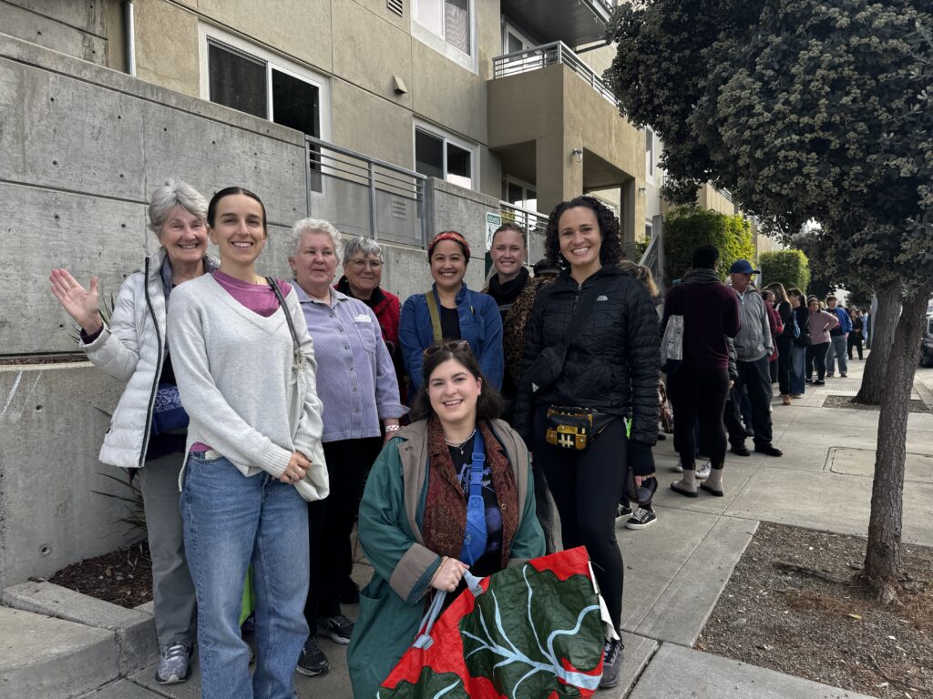 A long line forms on a sidewalk outside an apartment building, with several women up front smiling for the camera.