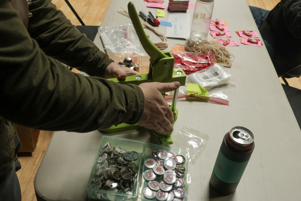 Hands work a green button-maker. To the right of the gadget is a box of button parts. The the left are rubber bands and red whistles rubberbanded to pink cards.