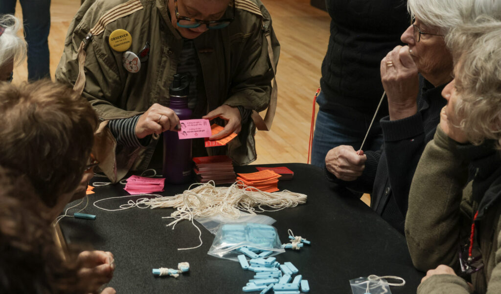 Hands hold a pink card at a table where three women sit looking at the card. In front of them, a table holds blue plastic whistles, string and orange and pink cards.