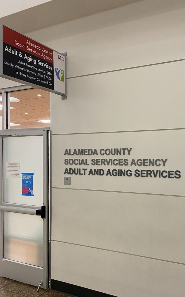 To the right of a doorway, letters engraved in black on a white stone wall identify the room as Alameda County Social Services Agency Adult and Aging Services office.